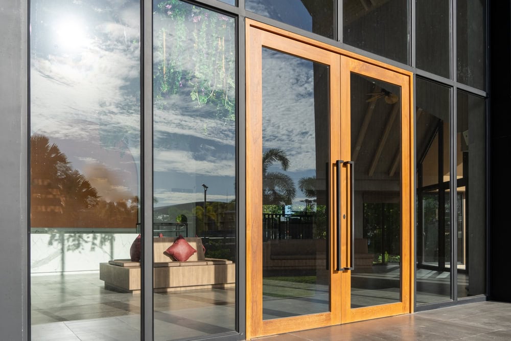 Modern glass building entrance with large wooden double doors and metal handles; outdoor scene reflected in the glass, showing FL blue sky and clouds. Interior features red cushions. Expert glass repair Pinellas County available for all needs.