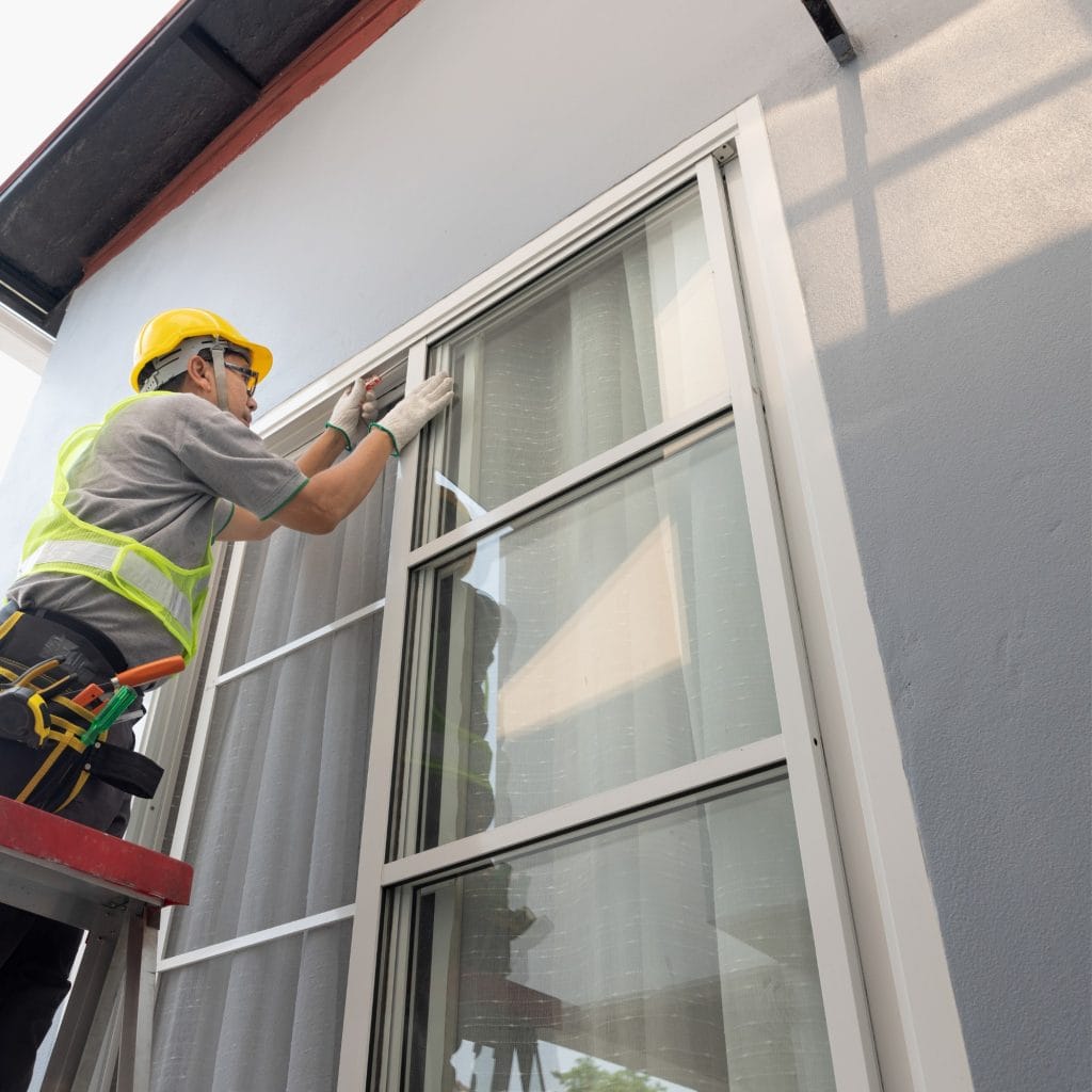 A construction worker in a yellow hard hat and safety vest installs or repairs a window on a gray building, standing on a ladder and using tools—typical of glass repair Pinellas County, FL projects.