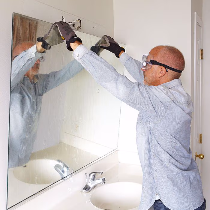 A person wearing safety goggles and gloves is carefully removing a large bathroom mirror above a sink, using a tool to detach it—mirroring the precision seen in commercial glass door repair Pinellas County, FL.