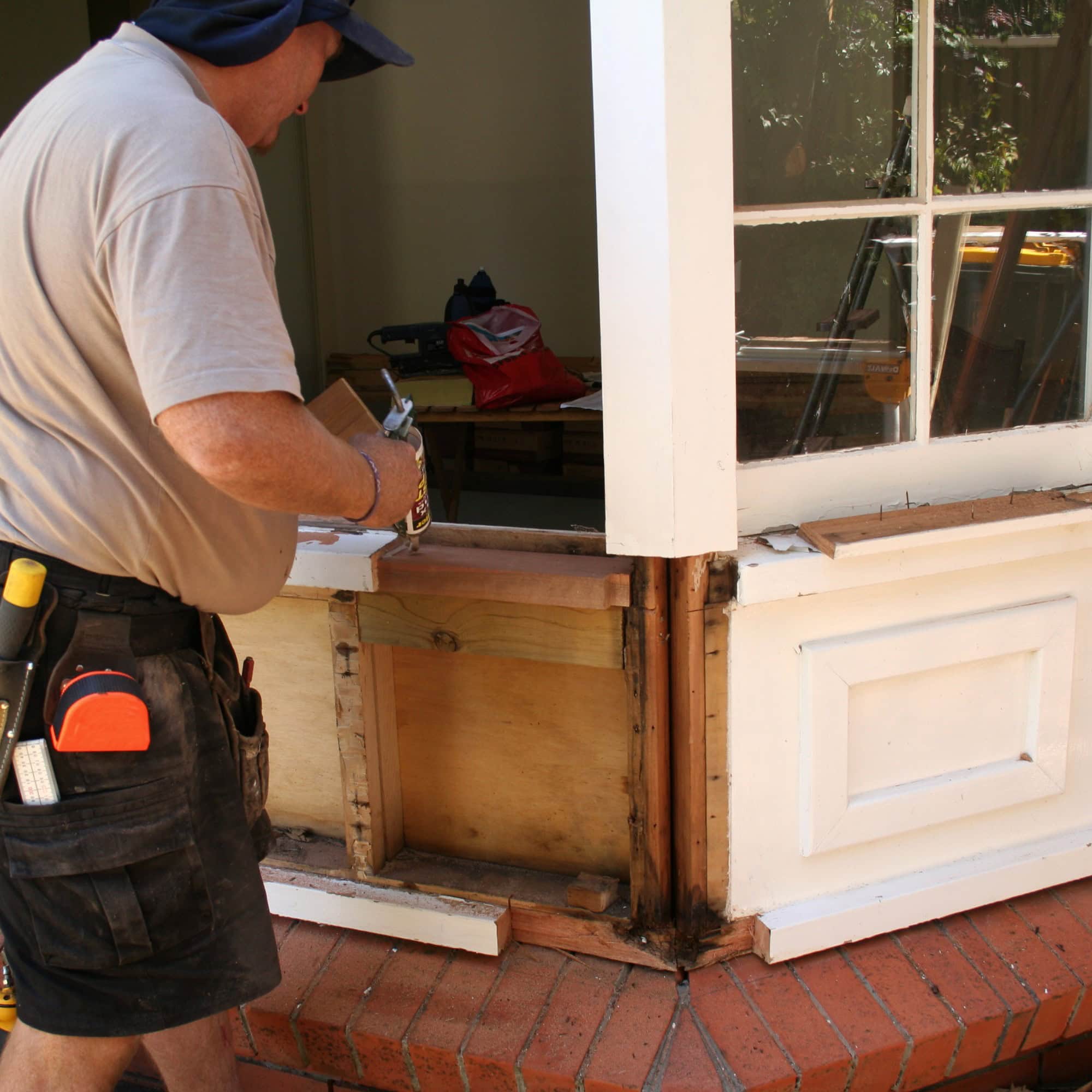A person wearing a hat and tool belt is repairing a wooden window frame on a brick porch in FL, applying material to exposed wood and damaged areas beneath a large window—perfect for glass repair Pinellas County projects.