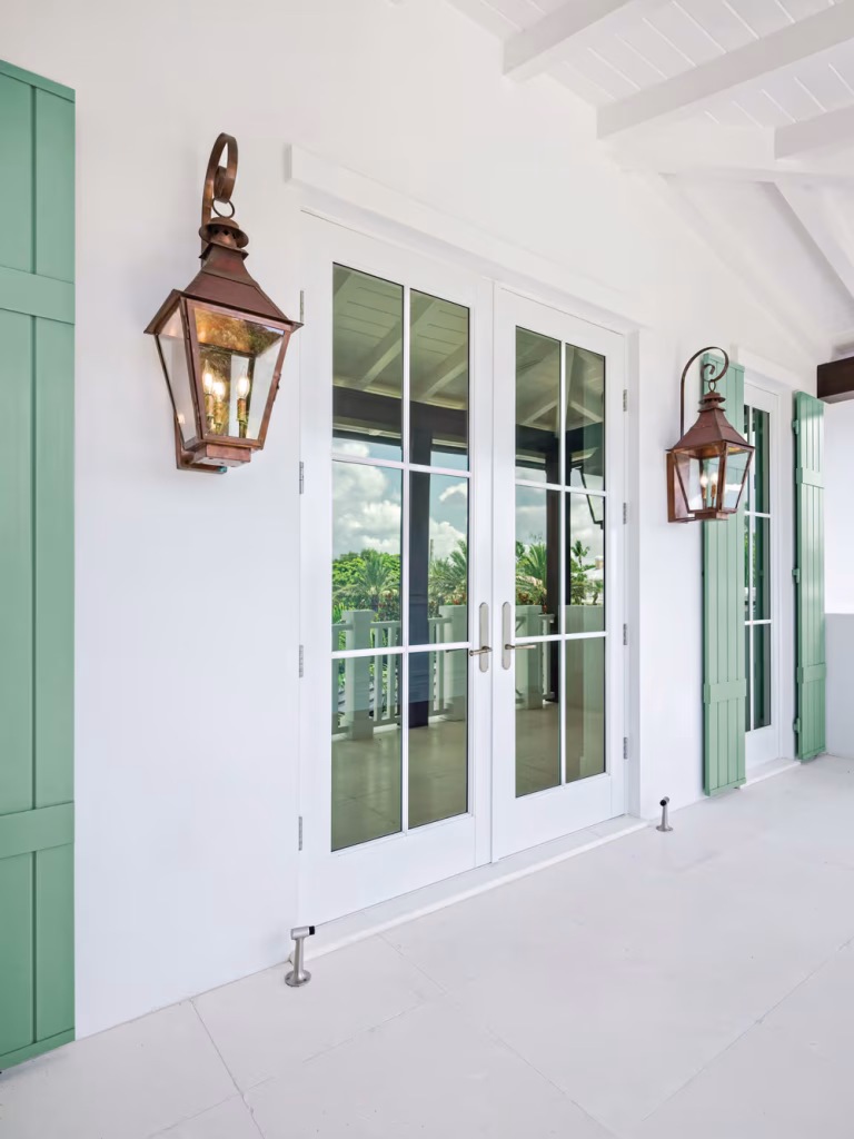 A pair of glass French doors with white frames open onto a balcony, flanked by green shutters and large copper lantern-style wall lights on a white exterior wall. For expert glass repair Pinellas County, FL residents trust, the trees and sky reflect in flawless panes.